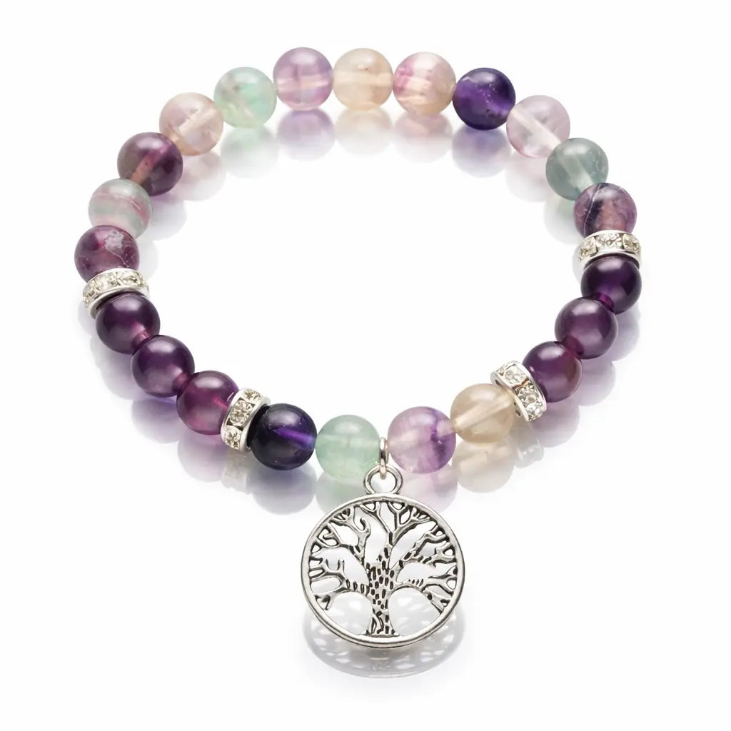 Un bracelet en pierres naturelles de fluorite, alternant des perles rondes multicolores (violet, vert, translucide), orné d'un pendentif Arbre de Vie en métal argenté et de perles de strass, présenté sur un fond blanc réfléchissant.