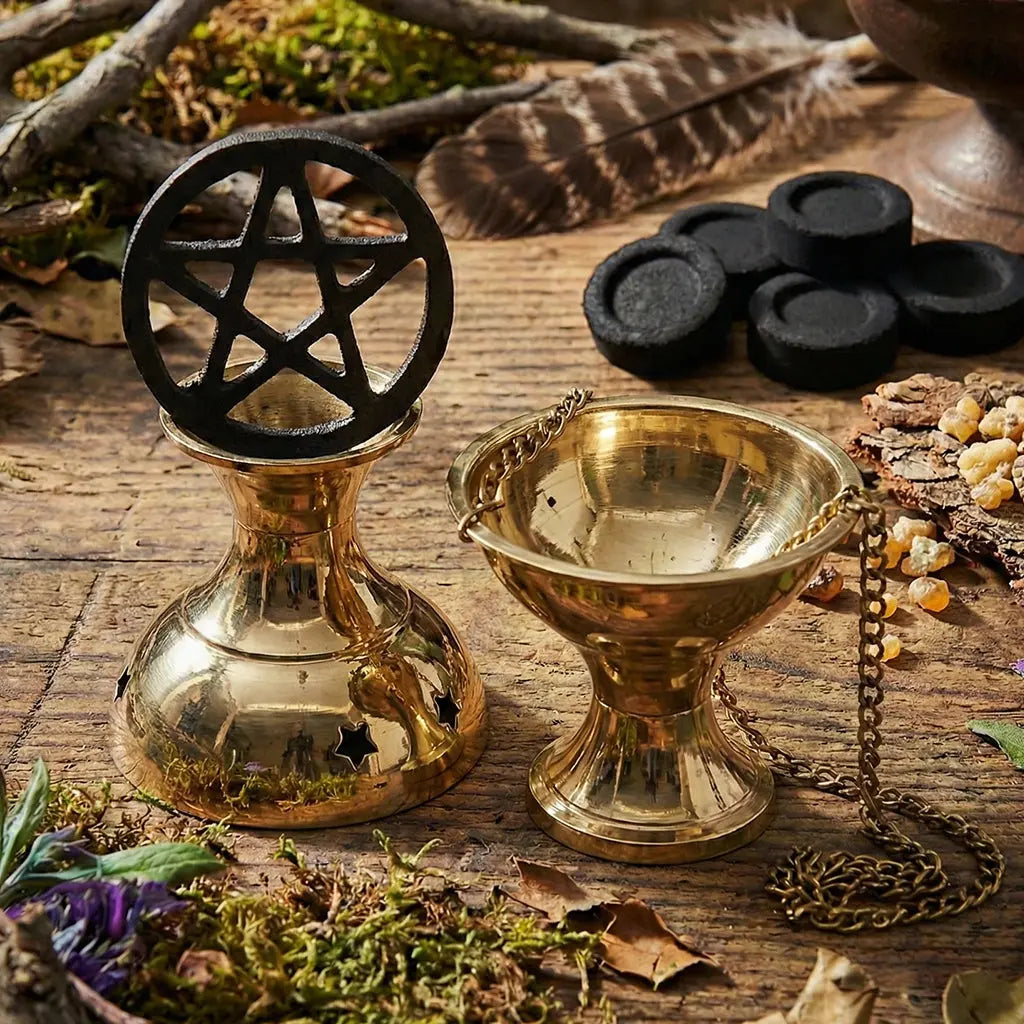 Brûle-encens/encensoir doré en laiton avec chaînette, posé sur une table en bois, accompagné d’un support avec symbole pentagramme et de charbons noirs, plumes et résine d’encens en arrière-plan.