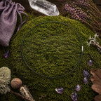 Câble gainé noir circulaire avec fermoir argenté, posé sur une butte de mousse avec une pointe de cristal, une pochette violette et des pierres d'améthyste.