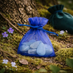 Petite pochette en organza bleu électrique contenant plusieurs pierres d'aigue-marine polies d'un bleu clair translucide, posée sur de la mousse au pied d'un arbre.