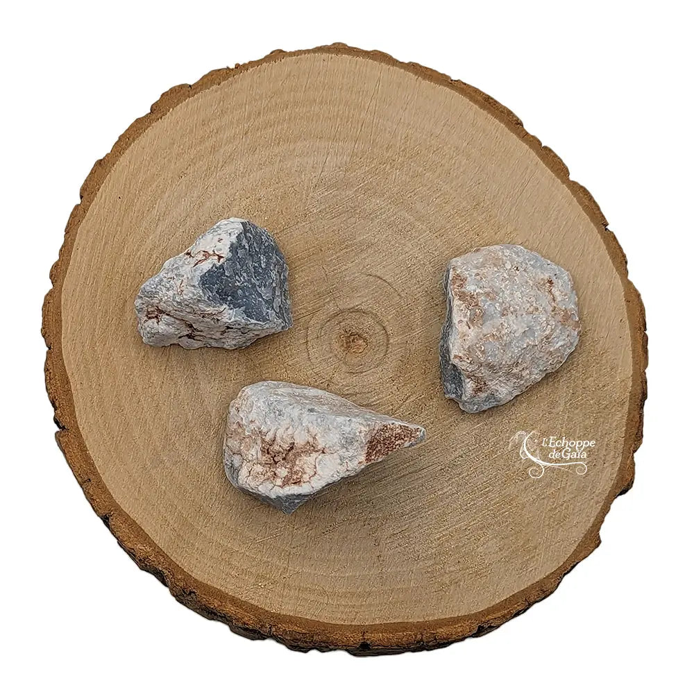 Trois morceaux d'angélite brute, présentant des teintes gris-bleu et des zones blanchâtres naturelles, disposés sur une tranche de bois clair.