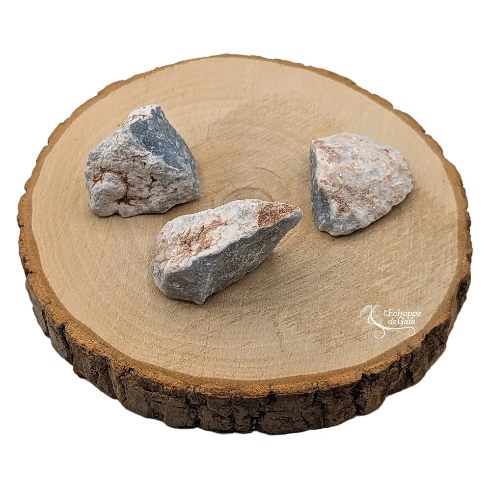 Trois morceaux d'angélite brute aux nuances bleu-gris et blanches, présentés sur une tranche de bois clair.