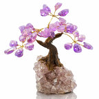 Une vue d'ensemble d'un petit arbre du bonheur montrant son tronc sculpté et ses feuilles en améthyste violette, solidement fixé sur une base de cristal de roche brut sur fond blanc.