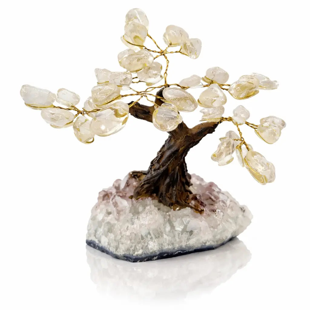 Un petit arbre du bonheur décoratif avec des feuilles en cristal de roche translucide et un tronc brun sculpté, fixé sur une base de cristal brut sur fond blanc avec reflet.