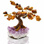 Un arbre du bonheur décoratif aux feuilles en pierres d'œil de tigre et branches dorées, s'élevant d'un socle en amas d'améthyste violette, présenté sur un fond blanc.