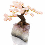 Un arbre du bonheur décoratif aux feuilles de quartz rose et tronc sculpté, s'élevant d'un socle en pointe d'améthyste brute, présenté sur un fond blanc.