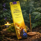 Boîte jaune lumineuse d'encens Saint Gabriel Archange Aromatika illustrée de l'Annonciation, posée sur une branche moussue avec une bougie et des copeaux de santal.