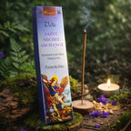 Boîte bleue d'encens Saint Michel Archange Aromatika illustrée du Prince de la Milice Céleste, posée sur une souche moussue en forêt avec un bâtonnet fumant, une bougie et des fleurs de violette.