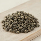 Gros plan sur un monticule de grains de résine d'Encens Benjoin de Sumatra, montrant des morceaux irréguliers de couleur gris-brun foncé/terreux, sur une planche de bois clair.