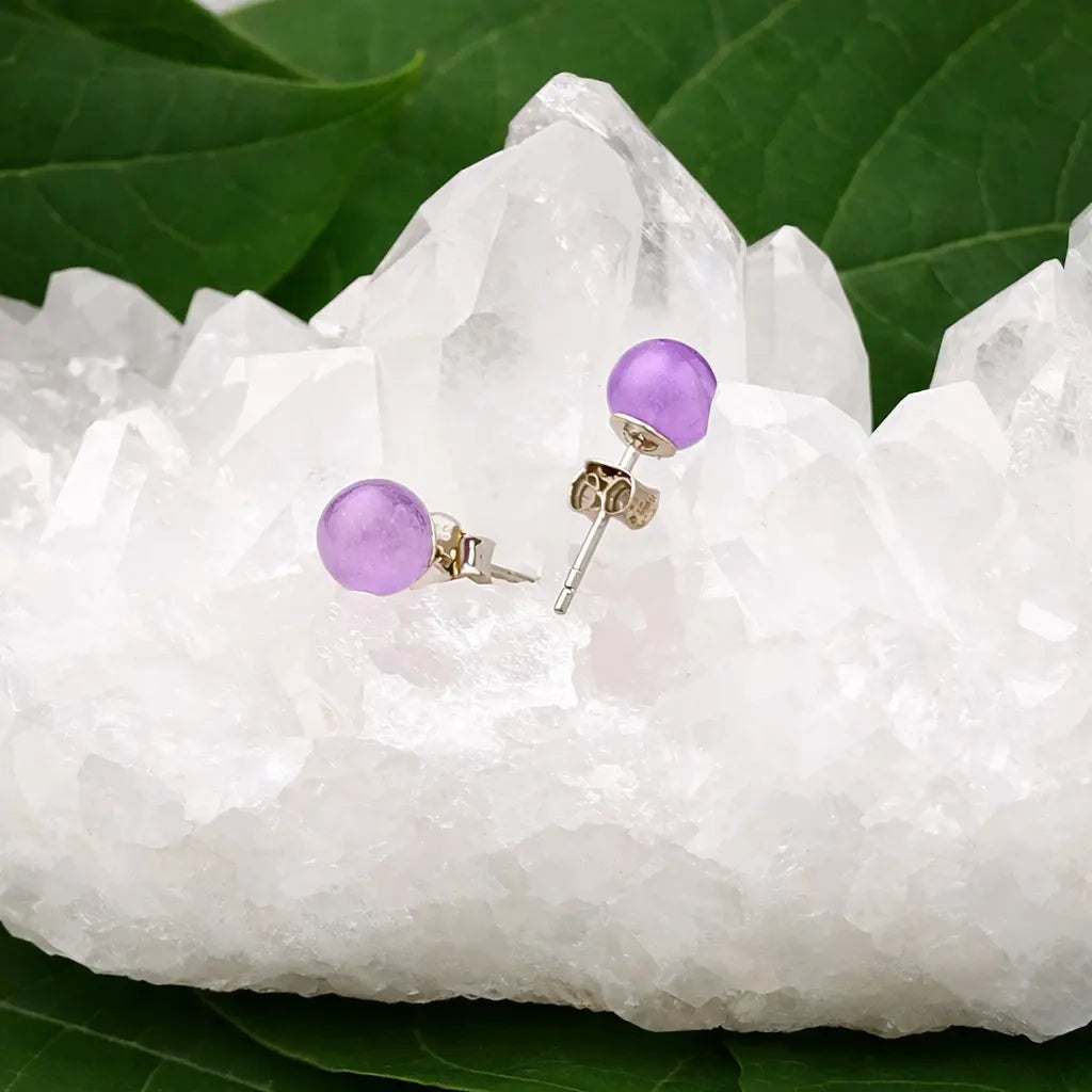 Une paire de boucles d'oreilles puces en améthyste violette, délicatement posées sur un amas de cristal de roche brut blanc, entouré de grandes feuilles vertes.