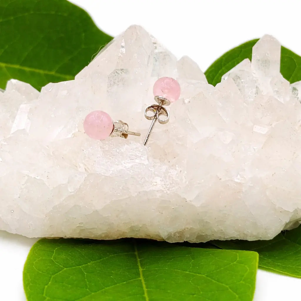 Une paire de boucles d'oreilles puces en quartz rose, délicatement posées sur un amas de cristal de roche brut décoré de quelques feuilles vertes.