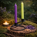 Un bougeoir double en fer forgé noir au design incurvé, supportant deux bougies colorées (violette et verte), mis en scène dans un décor naturel sur une tranche de bois entourée de mousse et de fleurs.
