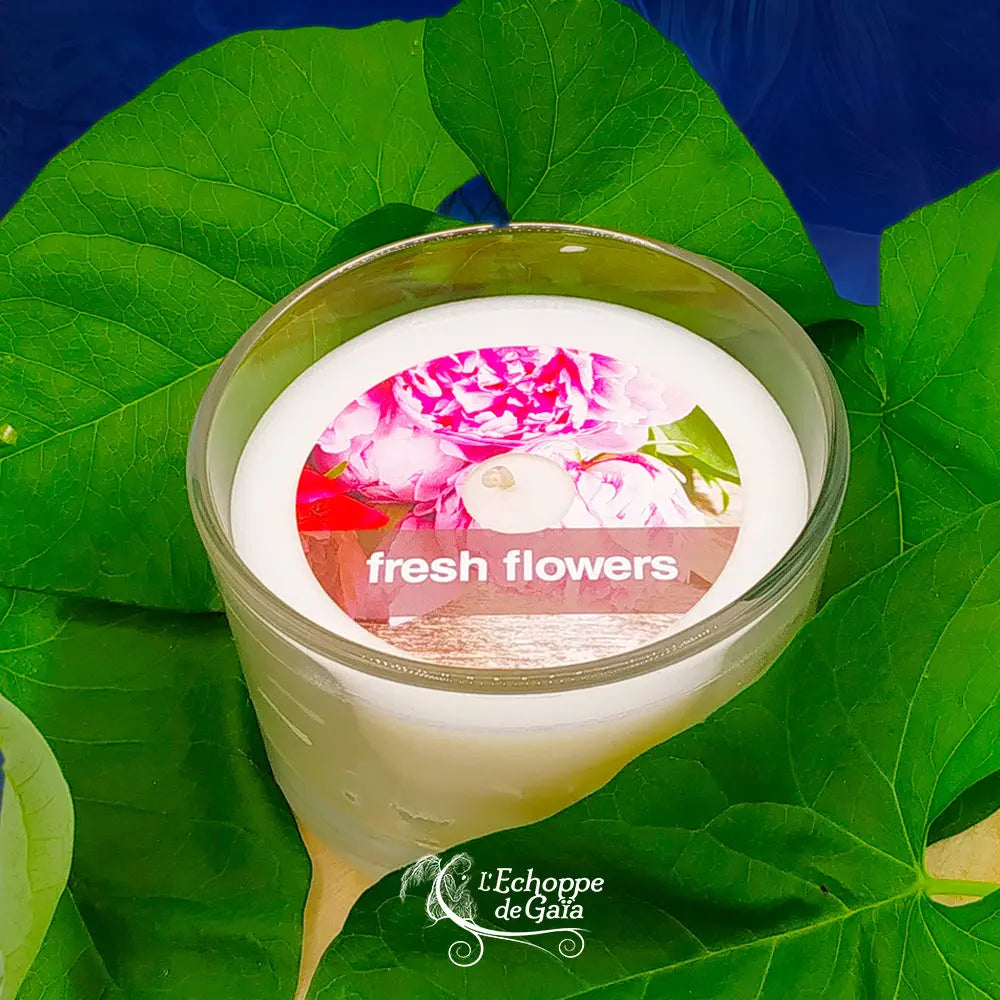 Bougie parfumée "fresh flowers" dans un pot en verre, présentée sur un lit de feuilles vertes.