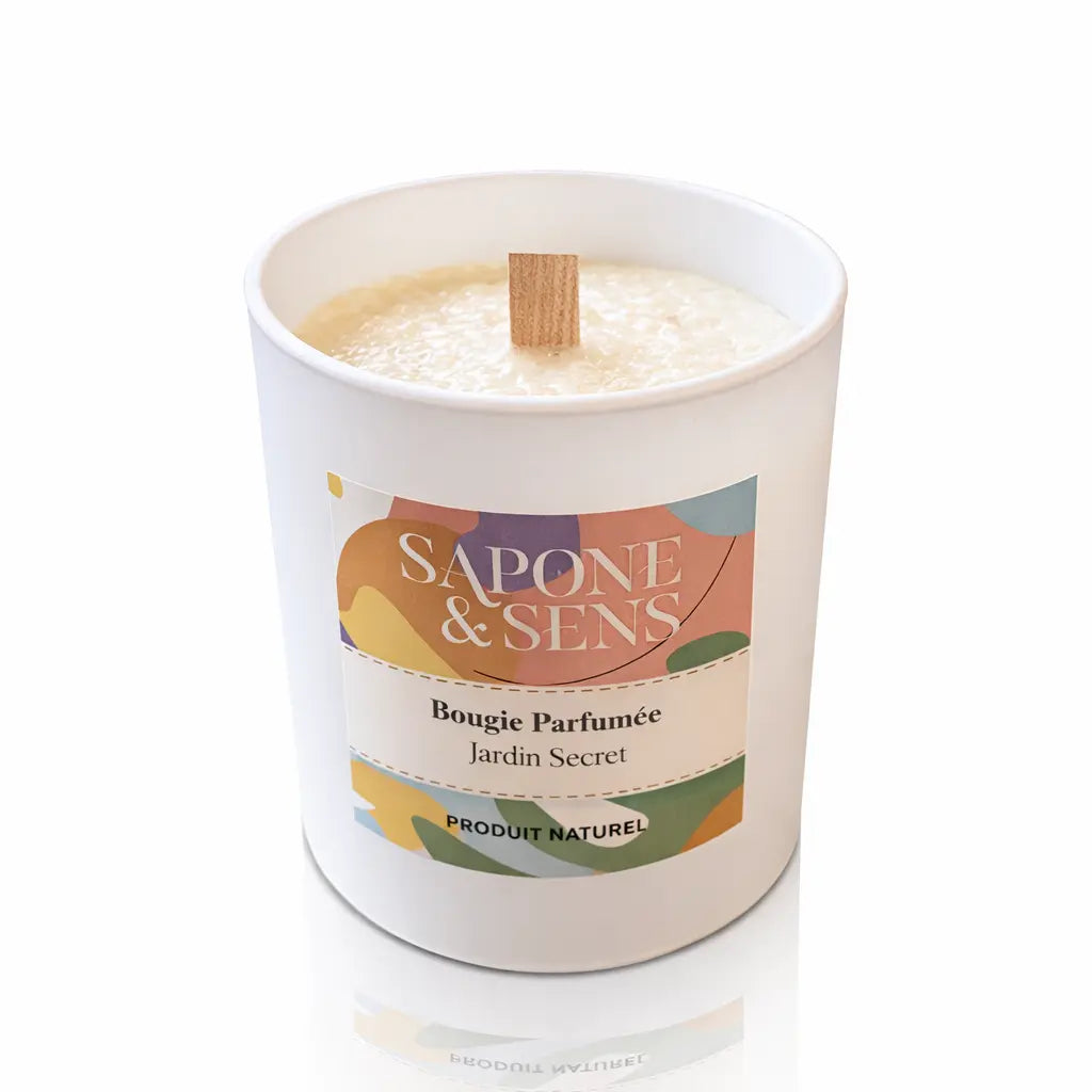 Une bougie parfumée naturelle « Jardin Secret » de la marque Sapone & Sens, présentée dans un pot en céramique blanche avec une mèche en bois, sur un fond blanc.