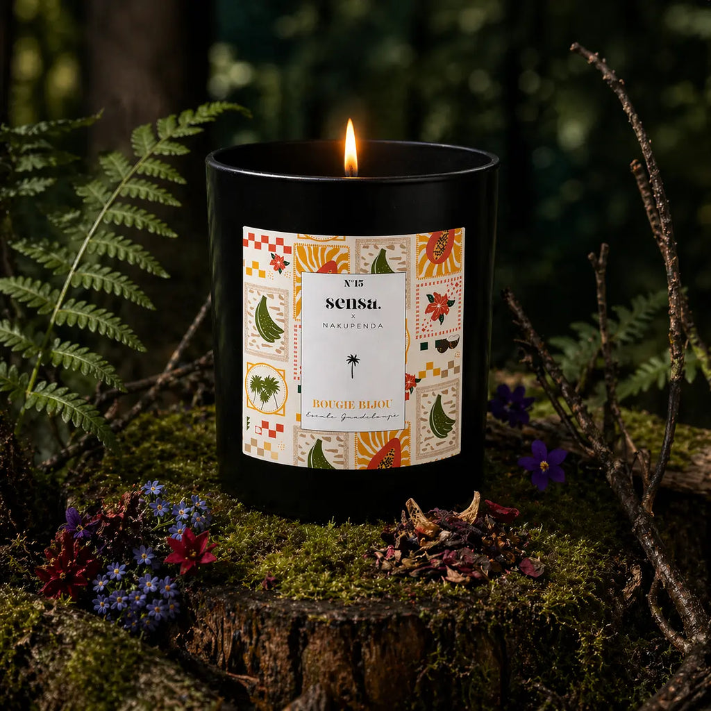 Bougie parfumée Eskalia Escale Guadeloupe à l'étiquette illustrée de motifs exotiques, allumée sur une souche moussue avec des fleurs violettes et des fougères.