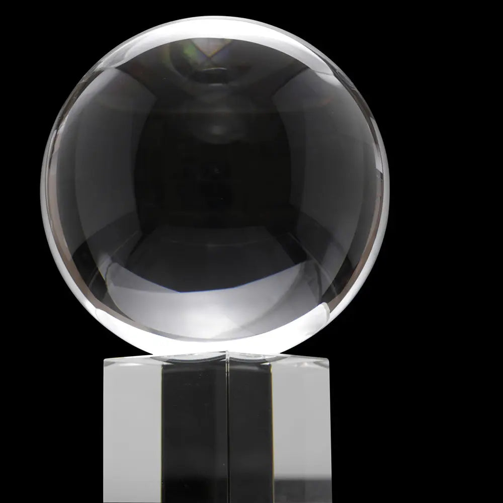 Boule de cristal très transparente, photographiée sur fond noir : la sphère capte de forts reflets lumineux et repose sur un socle cubique en verre/plexi clair.