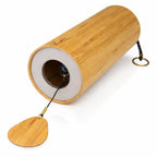Un carillon Koshi Ignis, composé d'un tube de résonance en bambou naturel avec son logo au point rouge, présenté couché sur un fond blanc pour laisser apparaître son mécanisme intérieur et son battant en bois.