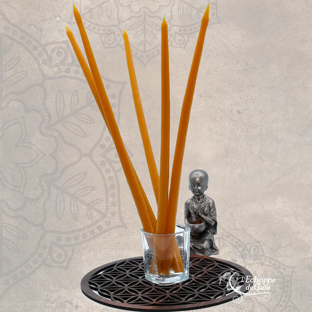 Cinq cierges fins en cire d'abeille jaune dans un verre, posés sur une Fleur de Vie avec une statuette de Bouddha.