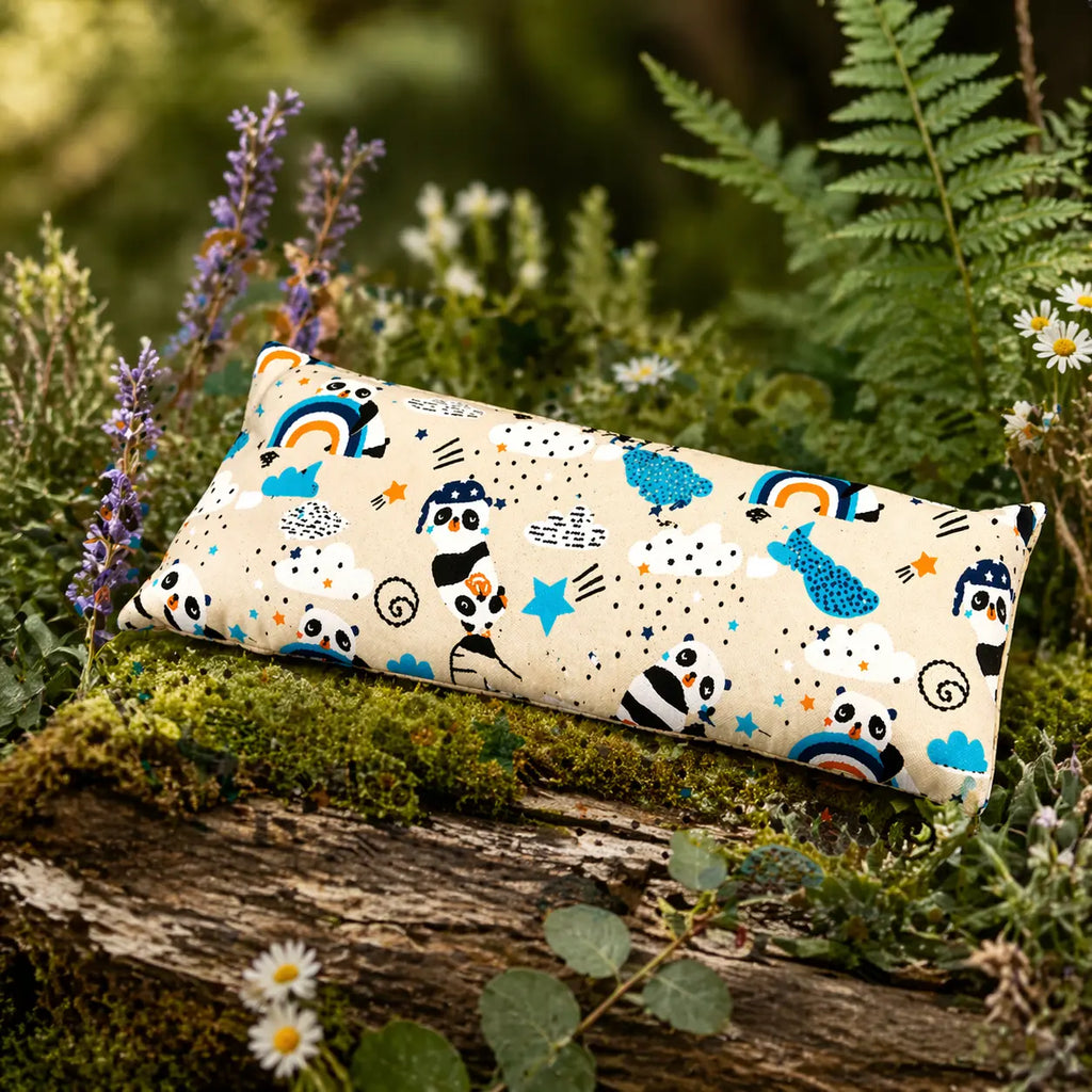 Coussin chauffant rectangulaire aux motifs de pandas, arcs-en-ciel et étoiles, posé sur un tronc d'arbre moussue en pleine nature, entouré de fleurs sauvages et de fougères.