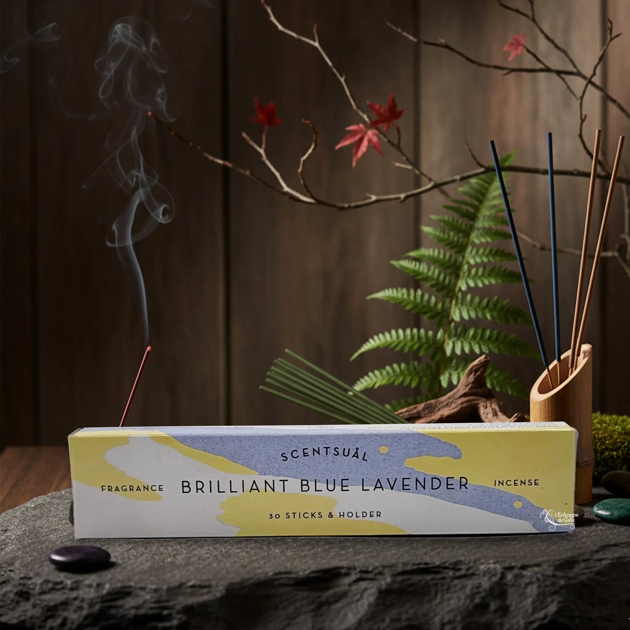 Boîte d'encens Scentsual Lavande Bleue posée sur une pierre naturelle, entourée de végétation et de bâtonnets d'encens dont la fumée s'élève avec douceur.