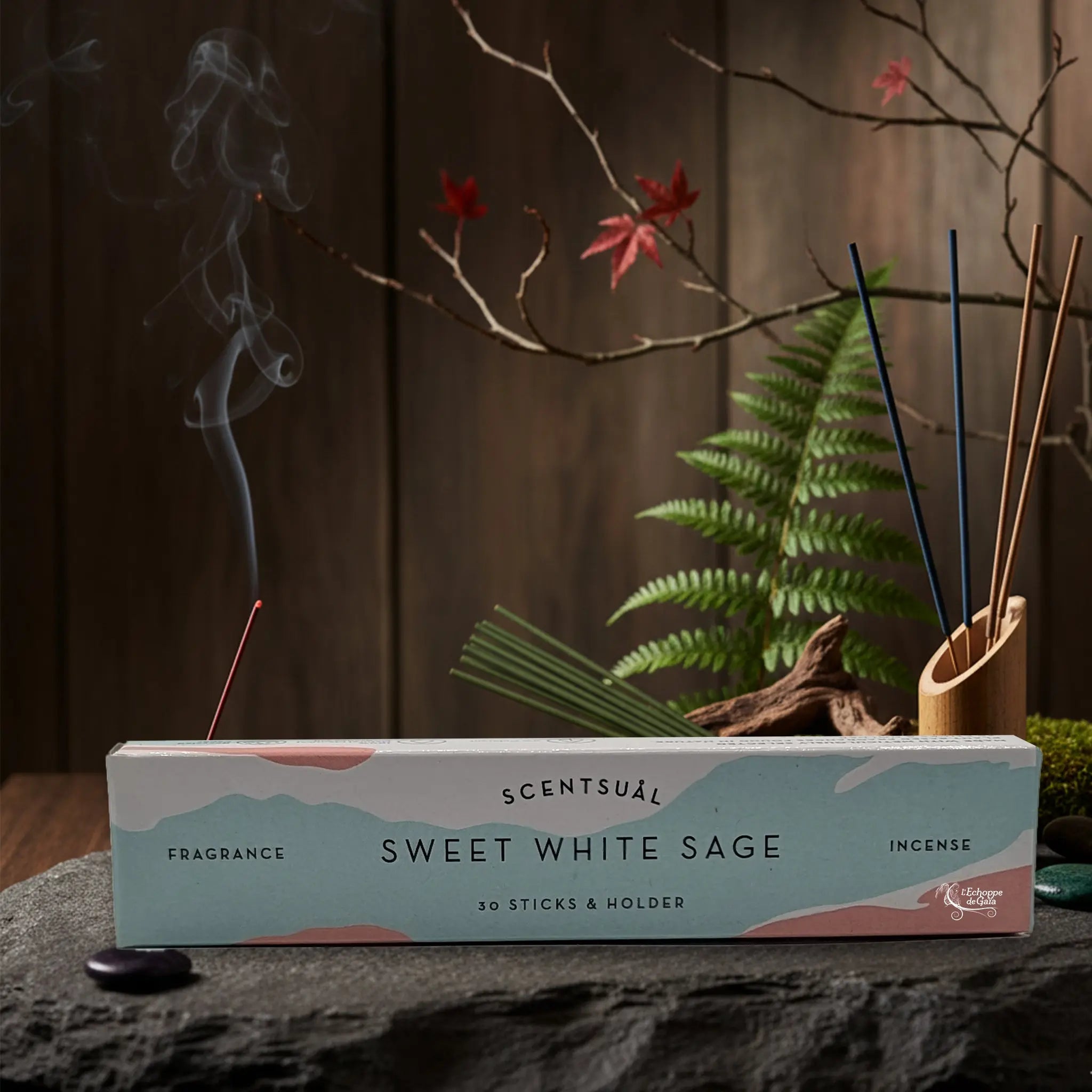 Boîte d'encens Scentsual Sweet White Sage posée sur un socle en pierre, accompagnée de bâtonnets d'encens bleus et marrons dans un décor naturel et zen.
