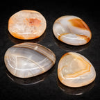 Quatre galets de cornaline polis aux formes lisses et organiques, révélant des motifs rubanés et des nuances orangées translucides, disposés sur un support noir brillant.