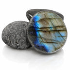 Labradorite - Demi-sphère, illustré au premier plan par la face plane et circulaire d'une pierre polie, révélant un éclat bleu électrique saisissant sur un fond minéral gris et strié, appuyée contre deux galets bruts.