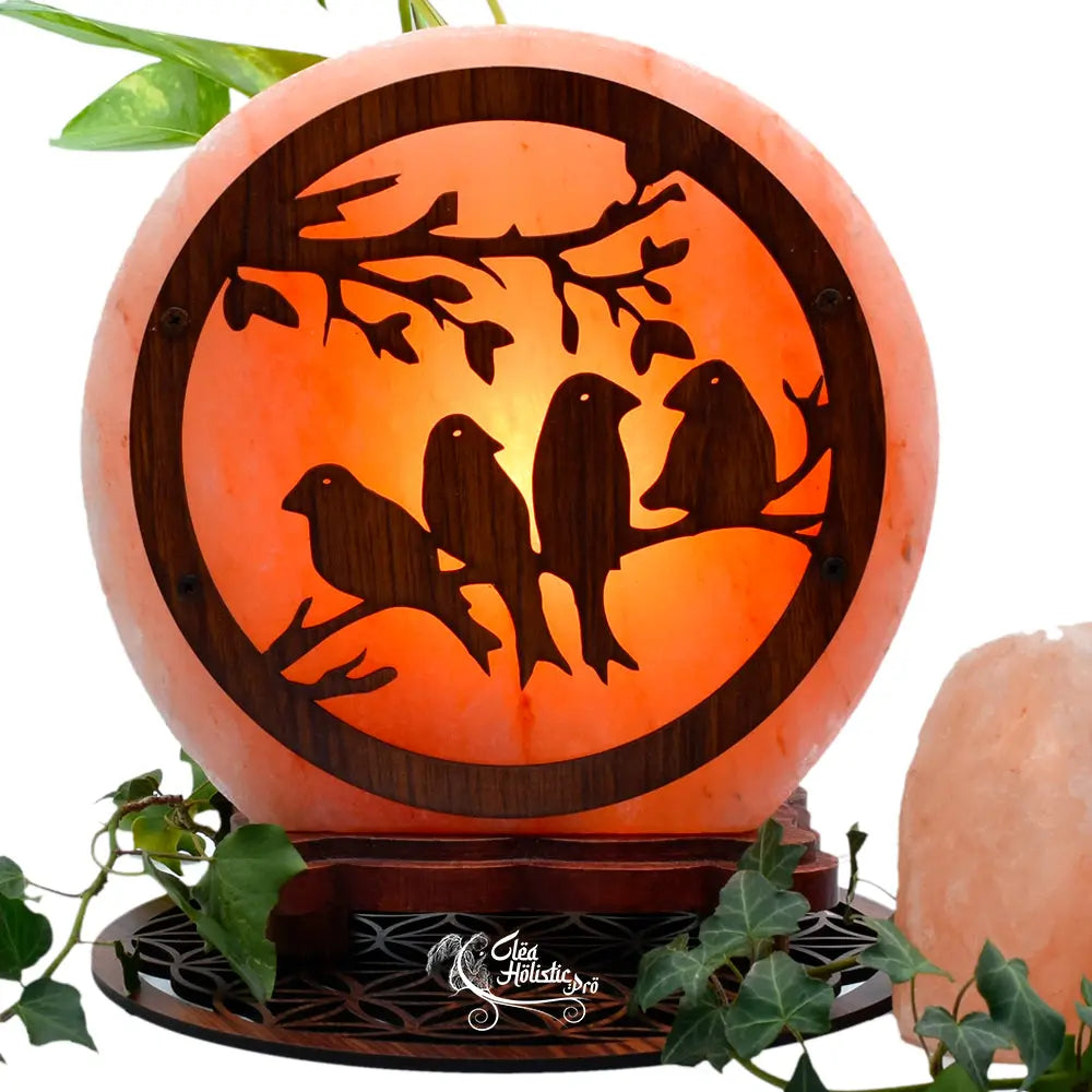 Lampe en sel de l'Himalaya de forme ronde, ornée d'un décor en bois représentant des oiseaux sur une branche et présentée parmi des feuilles de lierre.