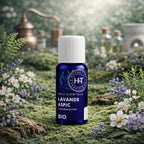 Flacon bleu d'huile essentielle de Lavande Aspic Herbes et Traditions posé sur un sol de mousse verte avec des fleurs de lavande et un alambic en cuivre.