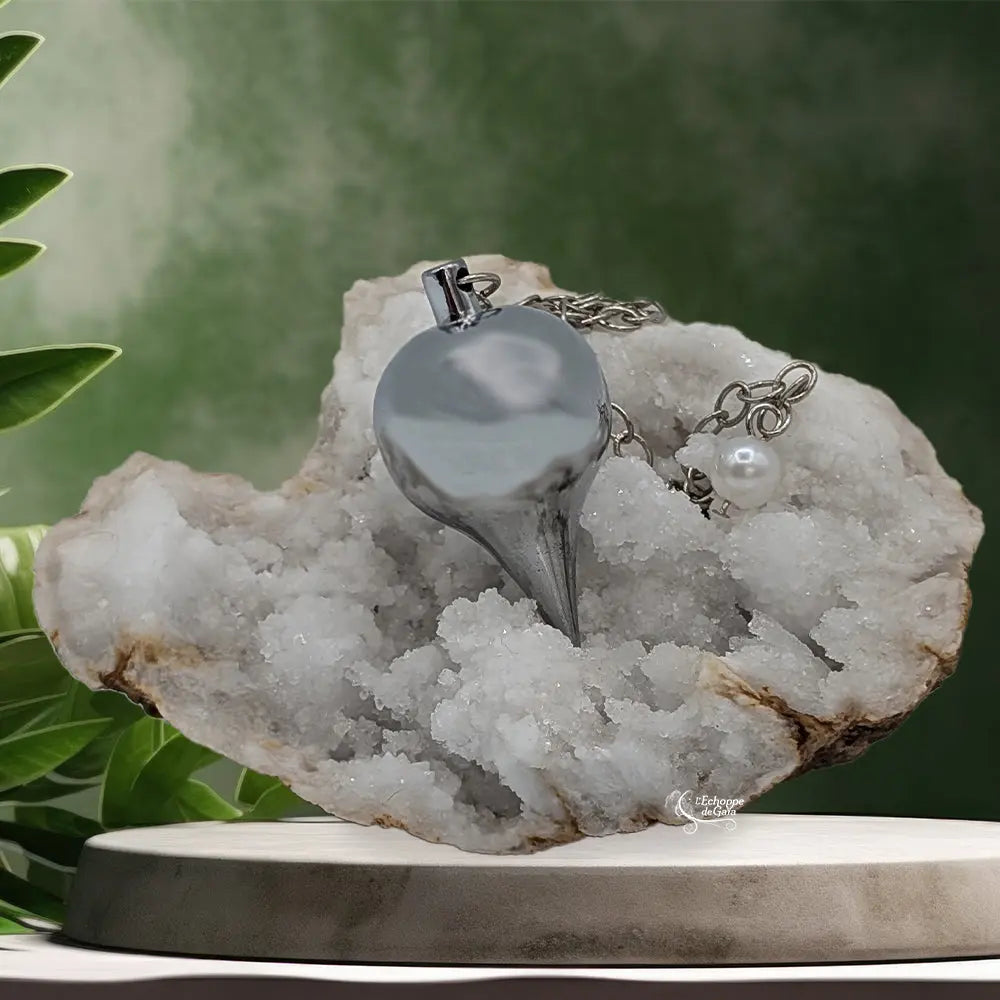 Pendule goutte en métal argenté brillant, posé sur une géode de cristaux blancs dans un décor zen.