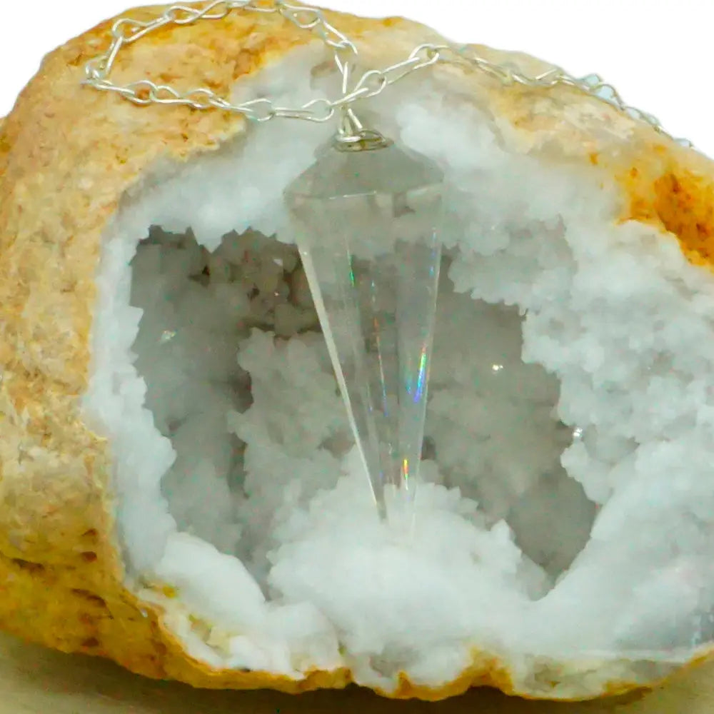 Pendule en cristal de roche taillé en cône à facettes avec chaîne en métal argenté suspendu devant une géode de cristaux blancs.