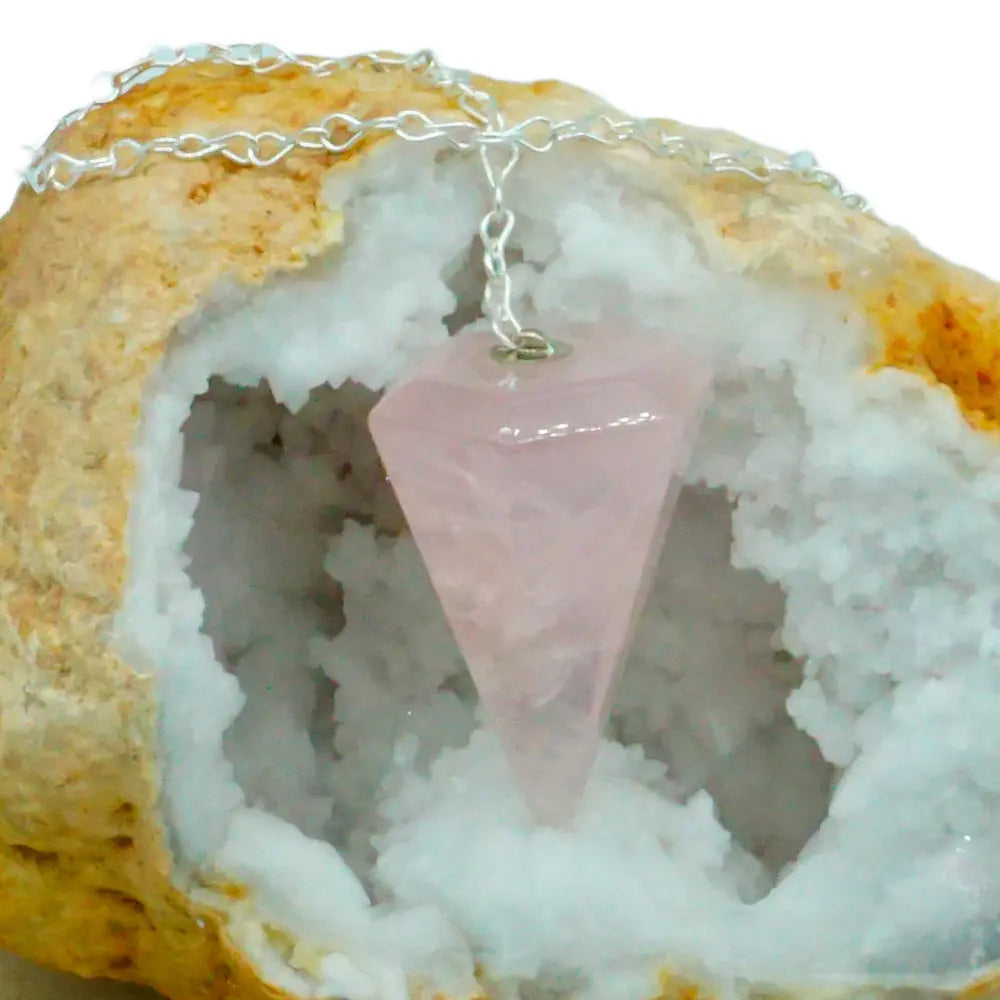 Pendule en quartz rose taillé en cône à facettes avec chaîne en métal argenté suspendu devant une géode de cristaux blancs.